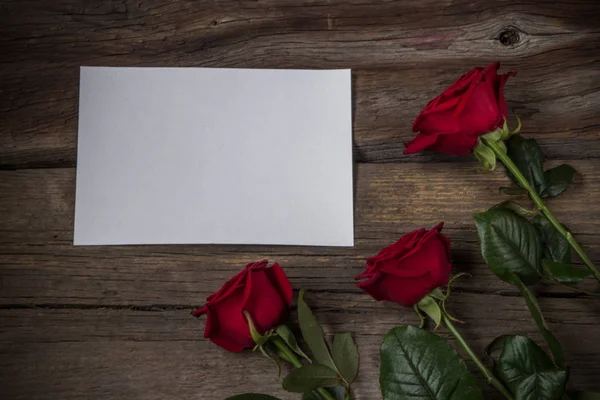 Rote Rosen und weißes leeres Papier auf hölzernem Hintergrund, Draufsicht Stockbild
