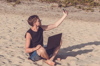 Dizüstü bilgisayar ve cep telefonu sahilde olan genç adam. Yapmak