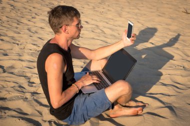 Dizüstü bilgisayar ve cep telefonu sahilde olan genç adam. Yapmak