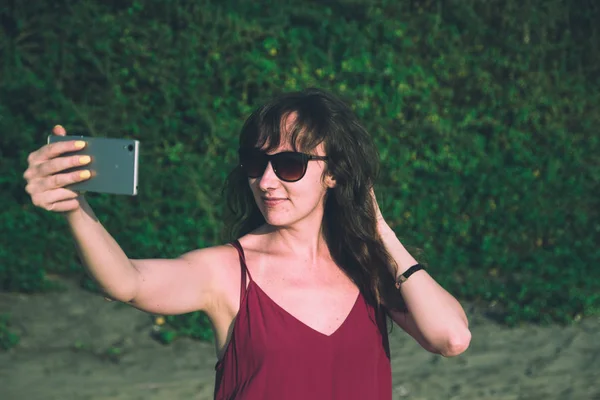 Wanita muda bergaun merah membuat foto selfie di pantai. Utara G — Stok Foto