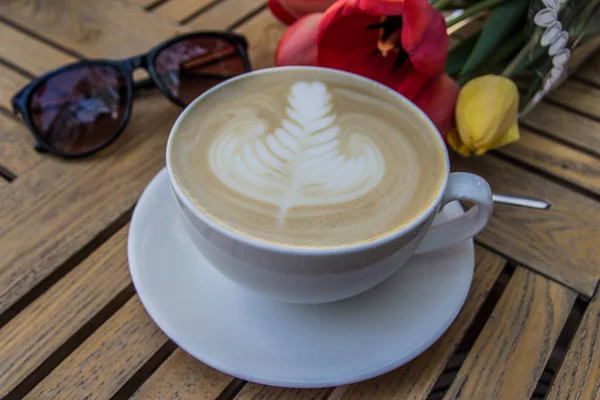 Cup of hot latte art coffee, glasses and tulips on wooden table — Stock Photo, Image