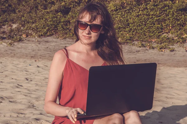 Young woman in red dress is working on laptop on the beach. Free — Stock Photo, Image