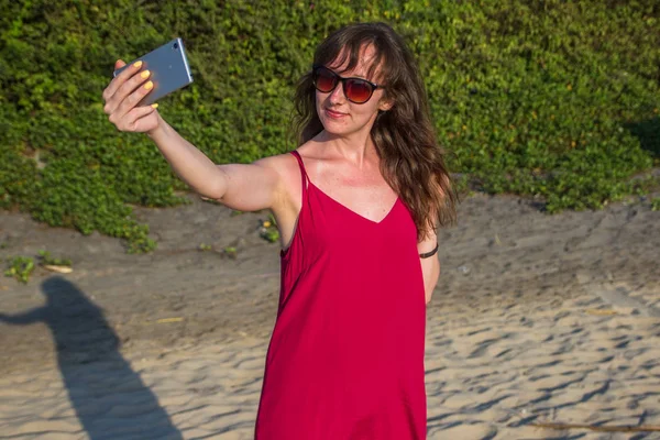 Молодая женщина в красном платье со смартфоном на пляже — стоковое фото