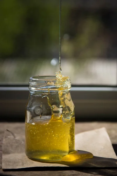 Βιολογικό αγνό μέλι σε βάζο στο περβάζι του παραθύρου. Ρεύμα του μελιού — Φωτογραφία Αρχείου