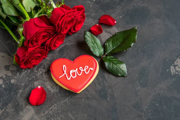 Κόκκινα τριαντάφυλλα και μπισκότο σε σχήμα καρδιάς με κείμενο αγάπη, σκούρο backgroun — Φωτογραφία Αρχείου