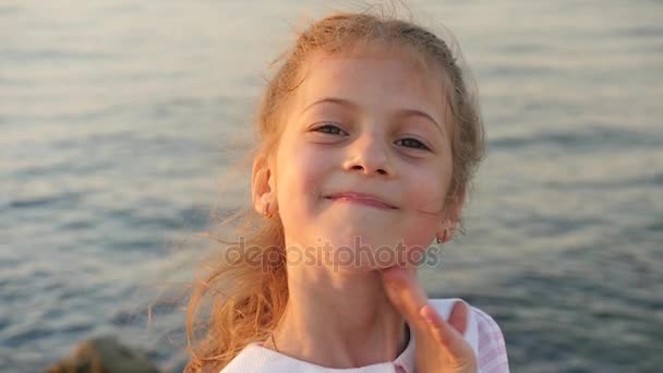 Красивая маленькая девочка на фоне заката моря улыбается показывая зубы — стоковое видео