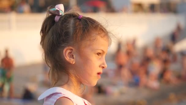 Bella sorridente bambina in piedi su una spiaggia sullo sfondo con le persone — Video Stock