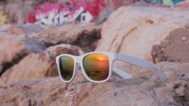 Sea zachód słońca odzwierciedlenie w okulary przeciwsłoneczne leżące na kamieniu — Wideo stockowe
