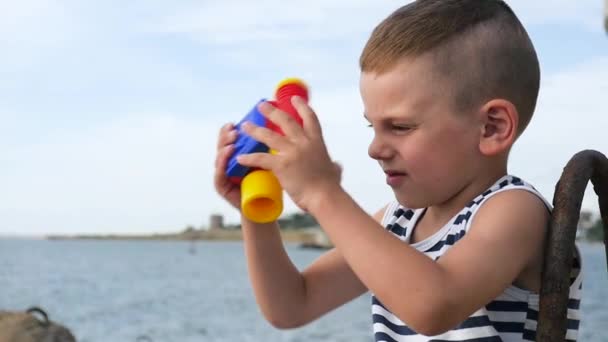 Ωραίο μικρό αγόρι στο ριγέ πουκάμισο κοιτάζοντας μέσα από κιάλια μπροστά στη θάλασσα — Αρχείο Βίντεο