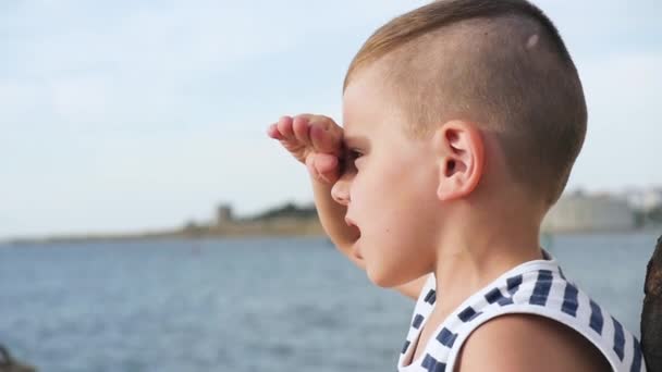 Маленька дитина в матросній сорочці, дивлячись на відстань на небі і морському фоні — стокове відео