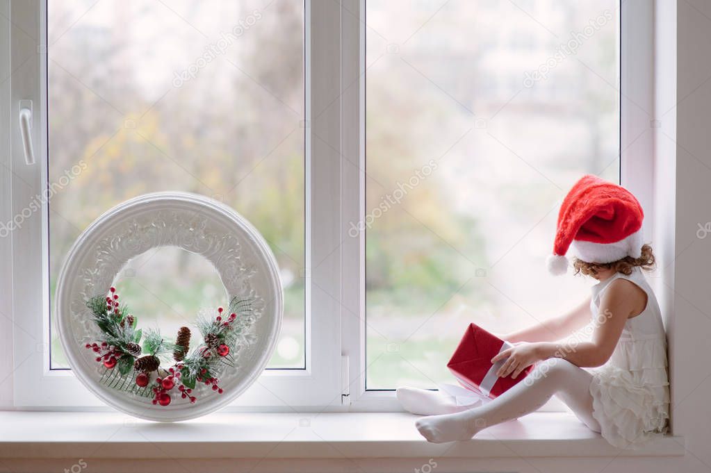 little girl in santa hat sitting by window with gifts and Christ