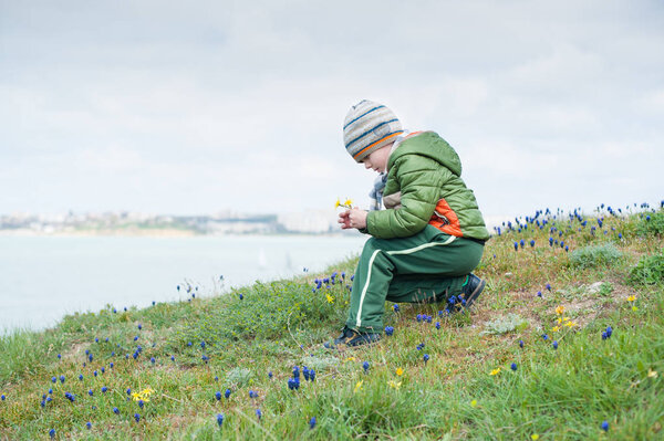 маленький ребенок в куртке и шляпе собирает весенние цветы на зеленом склоне на фоне моря
