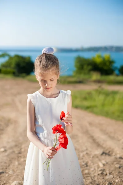 Pensive little girl wearing white dress with a bouquet of red poppies outdoors in spring — Stock Photo, Image