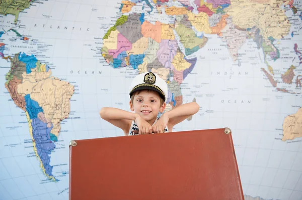 beautiful small boy in the captain\'s hat raises brown suitcase against background of the world map