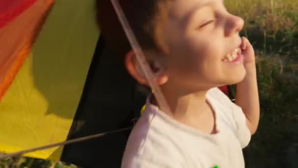 滑稽的小男孩与鬼脸在他的脸上拿着彩色风筝的手柄在他身后的夕阳夏天 — 图库视频影像