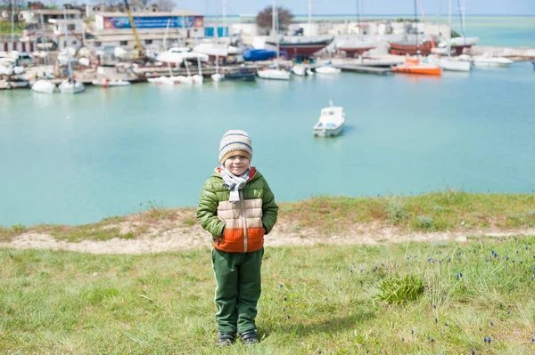 Gelukkig jongetje in warme jas en muts en sjaal op groene helling tegen de achtergrond van de zee baai met schepen in het vroege voorjaar — Stockfoto