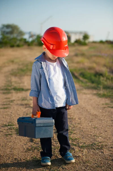 Грустный маленький рабочий парень в оранжевом шлеме повесил голову с футляром в одной руке, стоя на пути — стоковое фото
