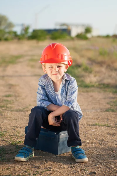 Lindo niño caucásico americano con casco naranja se sienta en la caja con herramientas después de un duro día de trabajo —  Fotos de Stock