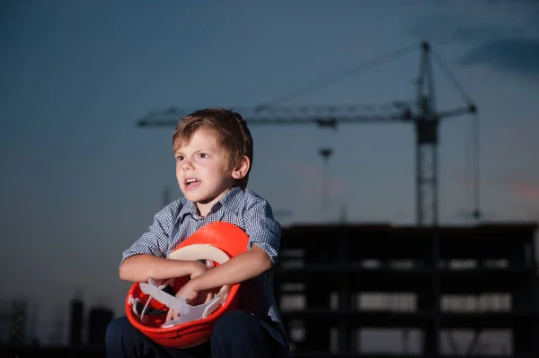 Кричащий маленький мальчик, сидящий с оранжевым шлемом в руках на фоне строительной площадки с краном в сумерках — стоковое фото