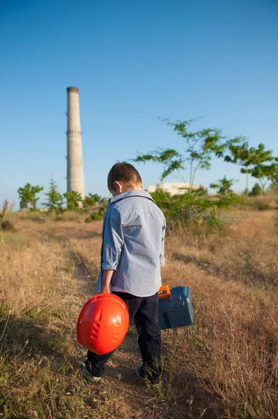 Triest Kaukasische jongetje met helm en gereedschap in handen lopen naar fabriek met pijp — Stockfoto