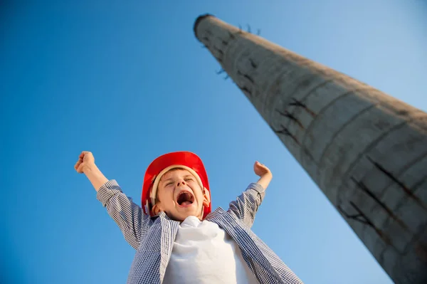 Juichende jongen in de helm aan de orde gesteld zijn handen tegen de blauwe hemel en fabriek pijp — Stockfoto