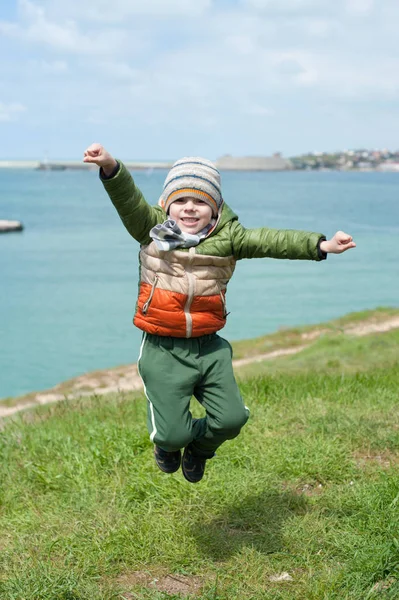 Смішна маленька дитина, одягнена в теплий шарф куртки і капелюх, стрибає на зеленому схилі на фоні морської бухти в холодну весну — стокове фото