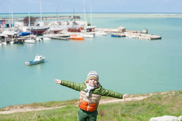 Ονειρική μικρό παιδί με σακάκι και καπέλο σε καταπράσινο λόφο εξάπλωση τα χέρια του σαν φτερά σε φόντο του λιμανιού στη θάλασσα με τις βάρκες στις αρχές της άνοιξης — Φωτογραφία Αρχείου