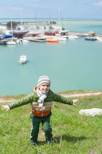 Felice bambino vestito con calda sciarpa giacca e cappello sul pendio verde sullo sfondo del porto marittimo con barche in primavera fredda — Foto Stock