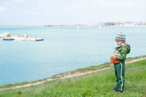 Cute pensive little boy in scarf jacket and hat on green grassy slope against sea with floating boats in springtime — Stock Photo, Image