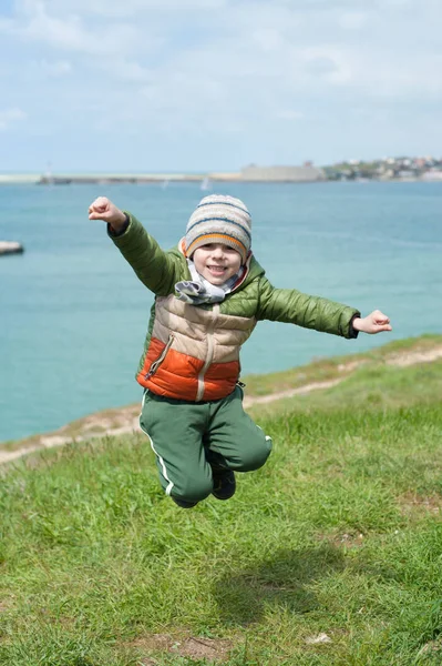 Grappige kleine jongen gekleed in warme jas, sjaal en muts springt op de groene helling tegen de achtergrond van de zee baai in het vroege voorjaar — Stockfoto