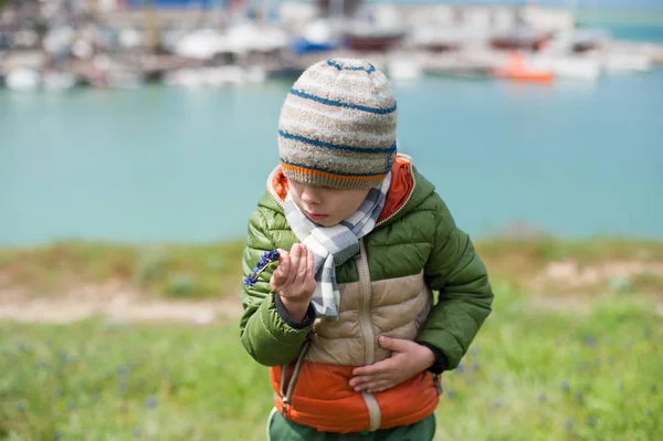 Curious little boy in jacket, scarf and hat looks surprised for Muscari flower in his hand on sea bay background in spring — Stock Photo, Image