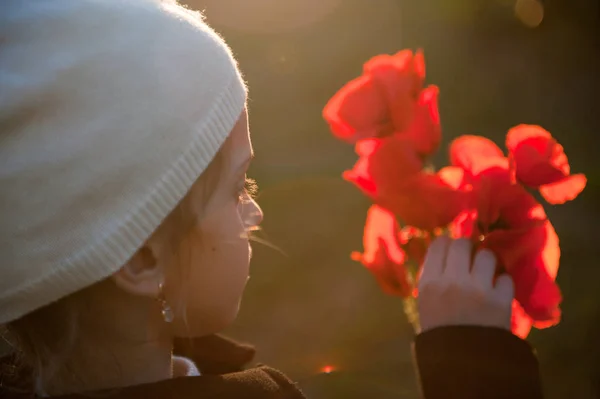 Linda menina em chapéu de lã branco tocando vermelho flores de papoula laranja em sua mão no pôr do sol de primavera — Fotografia de Stock