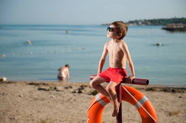 Cool piccolo ragazzo in pantaloncini rossi in occhiali da sole si siede sul palo con salvagente rosso appeso su di esso in spiaggia estiva — Foto Stock