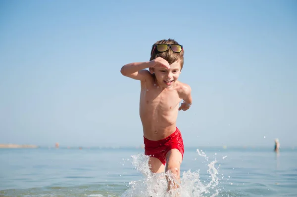Sano carino bambino felice indossando occhiali da sole e pantaloncini rossi in esecuzione in acqua dell'oceano con spruzzi in vacanza estiva — Foto Stock