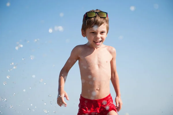 Felice sorridente carino bambino in occhiali da sole e pantaloncini rossi in acqua di mare con spruzzi in estate giornata di sole — Foto Stock