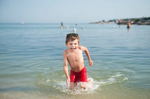 Felice ridere bambino in occhiali da sole e pantaloncini rossi in esecuzione in acqua di mare con spruzzi in giorno di sole estivo — Foto Stock