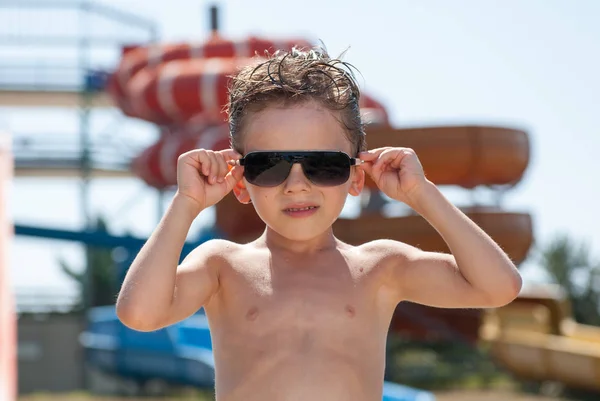Bellissimo bambino che indossa occhiali da sole alla moda nel parco acquatico con scivoli d'acqua in estate — Foto Stock
