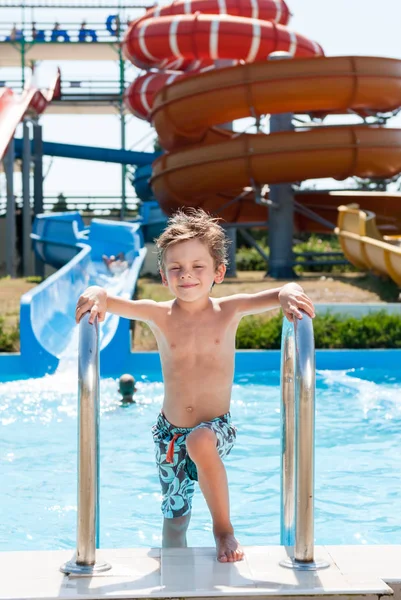 Felice sorridente bambino in pantaloncini in piedi vicino alla piscina nel parco acquatico con scivoli tubo d'acqua — Foto Stock
