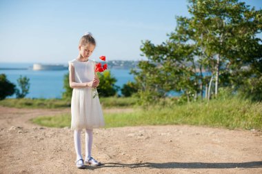 lonely thin little girl in white dress holding bouquet of red poppy standing on sea bay background clipart