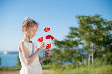cute little girl in white dress holding bouquet of red poppy standing on sea bay background clipart