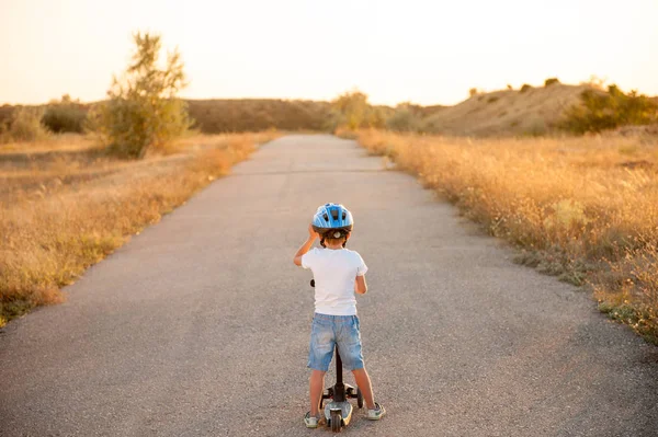 Eenzaam klein kind in de sport helm permanent op verlaten weg met scooter in zomer zonsondergang — Stockfoto