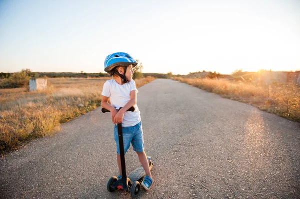 Gezonde kleine jongen in sport helm permanent op verlaten weg met scooter in warme zomer zonsondergang in de Verenigde Staten, Californië — Stockfoto