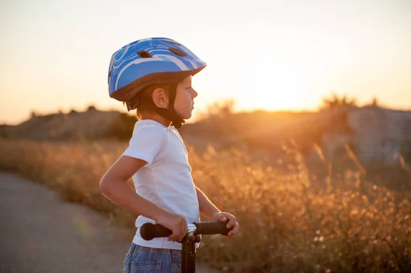 Pensieroso piccolo ragazzo in casco sportivo in piedi su strada abbandonata con scooter a guardare al caldo tramonto estivo — Foto Stock