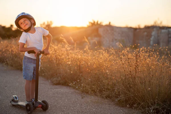 Glimlachend klein kind in de sport helm rijden op verlaten weg met scooter in de warme zomer zonsondergang — Stockfoto