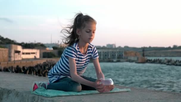 小さな女の子は Takes 小さな金魚鉢で 海ビューの背景都市桟橋に対して座っているし 小さな魚を見ています コンセプト レクリエーション — ストック動画