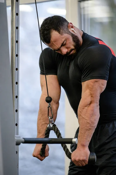 Strong healthy young bearded sport man in black sportswear and wireless headset during workout for muscle triceps pulling heavy weight using rope — ストック写真