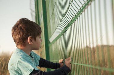 poor dirty little caucasian boy refugee standing near state border fence holding it with hands with hope of help and seeking for refuge