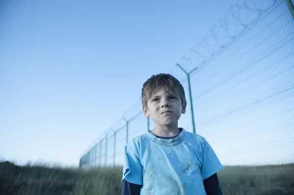 Portrait of poor orphan refugee prisoner migrant kid in dirty clothes with sad face near state border with high fence with barbed razor wire in concentration camp — Stockfoto