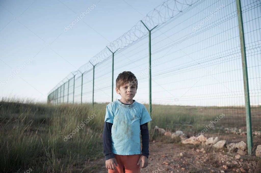 poor little caucasian refugee boy in dirty clothes standing near high fence with barbed razor wire at state border