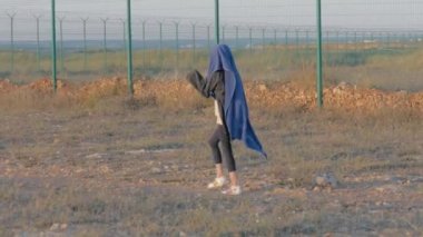 Evsiz çocuk koruma alanının yakınında yürüyor. Sıska mülteci kız başı örtülü yorgan demir çit eyalet sınırı boyunca uzanıyor. kavram: savaş sonrası ve siyasi mülteciler.